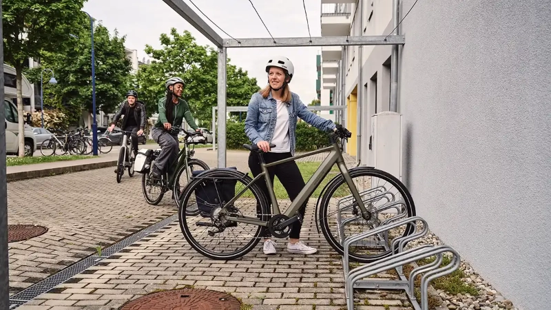 Eine Frau in blauer Jeansjacke parkt ihr grünes E-Bike, eine weitere Frau und ein Mann fahren den Fahrradstellplatz an