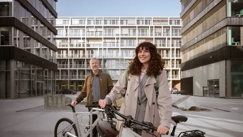 JobRadlerin steht mit Fahrrad abfahrbereit auf einem Firmencampus, im Hintergrund Kollege mit Fahrrad JobRadlerin steht mit Fahrrad abfahrbereit auf einem Firmencampus, im Hintergrund Kollege mit Fahrrad