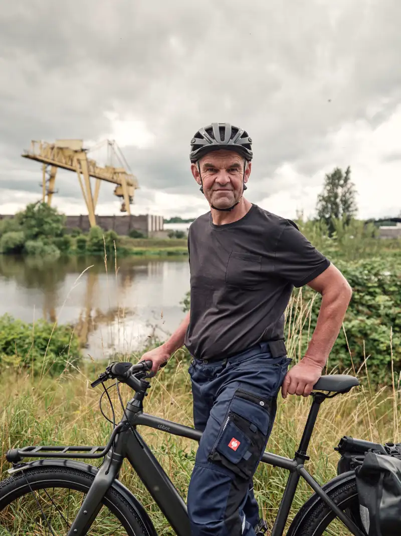 JobRadler in Arbeitskleidung im Grünen mit Kran im Hintergrund JobRadler in Arbeitskleidung im Grünen mit Kran im Hintergrund