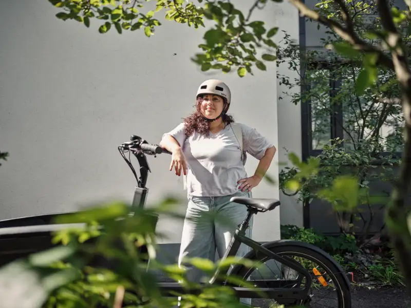 Frau mit Locken lehnt am JobRad, im Vordergrund Pflanzen Frau mit Locken lehnt am JobRad, im Vordergrund Pflanzen