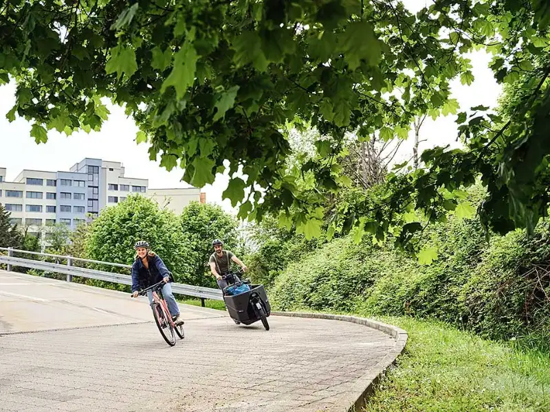 Eine Frau auf einem Stadtrad und ein Mann auf einem Lastenrad fahren gemeinsam durch eine Kurve Eine Frau auf einem Stadtrad und ein Mann auf einem Lastenrad fahren gemeinsam durch eine Kurve
