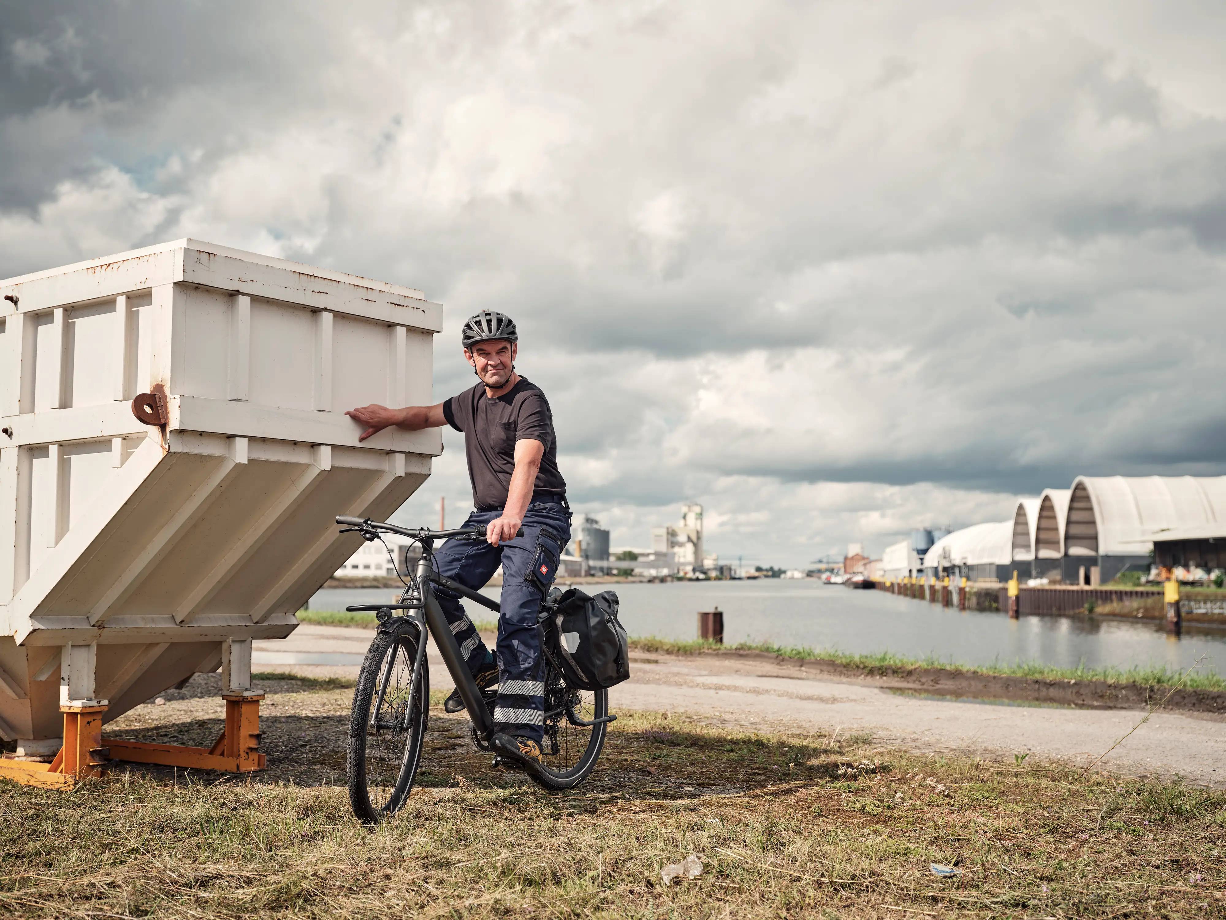 JobRadler in Arbeitshose lehnt an Container, hinter ihm Wasser JobRadler in Arbeitshose lehnt an Container, hinter ihm Wasser