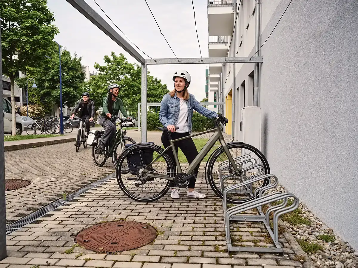 Eine Frau in blauer Jeansjacke parkt ihr grünes E-Bike, eine weitere Frau und ein Mann fahren den Fahrradstellplatz an
